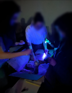 Trois joueuses examinent une boite en carton contenant des bouteilles, à la lumière d'une petite lampe à UV