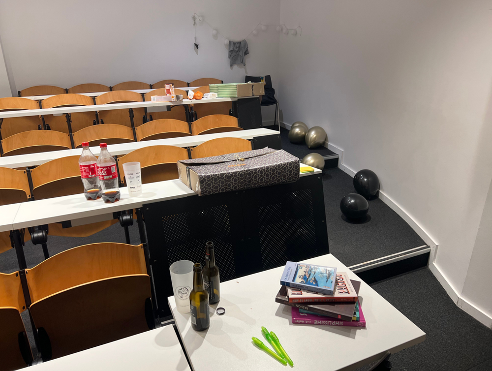 La salle d'escape game, éclairée normalement. Il y a plusieurs rangées de tables. Sur chaque rangée se trouve un ensemble d'objets correspondant chaun à une énigme.