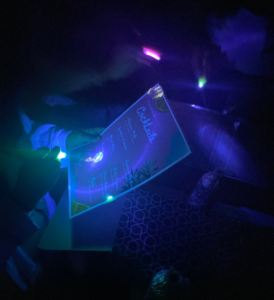 Une main tient un papier sur lequel est écrit une liste de cocktails, éclairé par une petite lampe à UV
