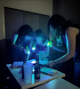 Trois joueuses examinent une feuille de papier à la lumière de petites lampes UV. Autour, sur la table, se trouvent des livres, des bouteilles de bière et un gobelet en plastique.